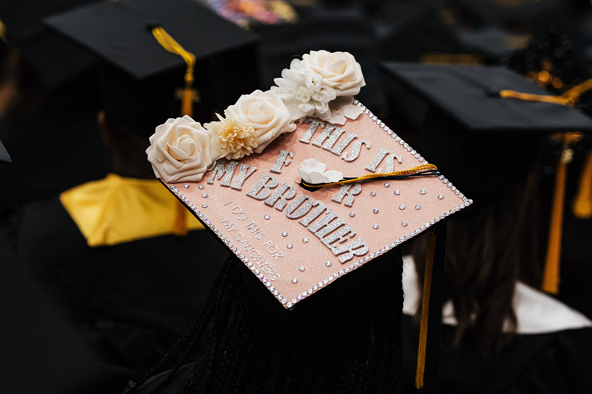 FLINT_Winter Commencement Caps April 2026_0051 Graduation cap decorated with white flowers and silver letters reading "This is for my brother, I do this for my daughter."