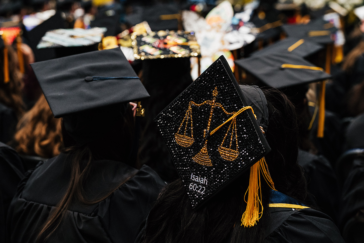 FLINT_Winter Commencement Caps April 2026_0052 Graduates wearing black caps and gowns, one cap decorated with gold scales of justice and the text "Isaiah 60:22".