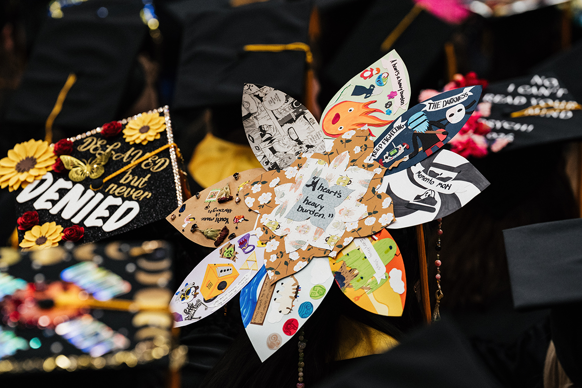 FLINT_Winter Commencement Caps April 2026_0055 Graduation cap decorated with a large flower made of paper petals, each petal featuring different drawings and messages, including "A heart's a heavy burden."