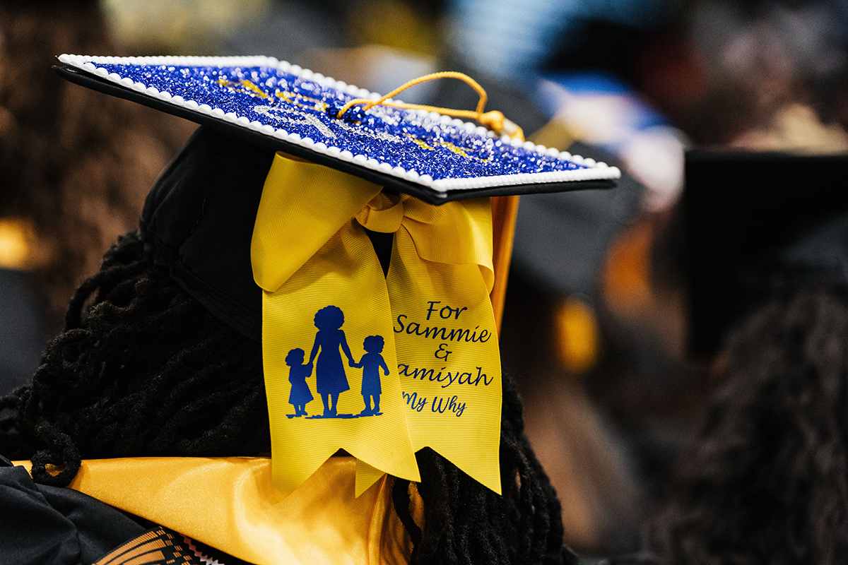 FLINT_Winter Commencement Caps April 2026_0060 Close-up of a decorated graduation cap with a yellow ribbon that reads "For Sammie & Amiyah My Why" and shows a silhouette of a woman holding hands with two children.