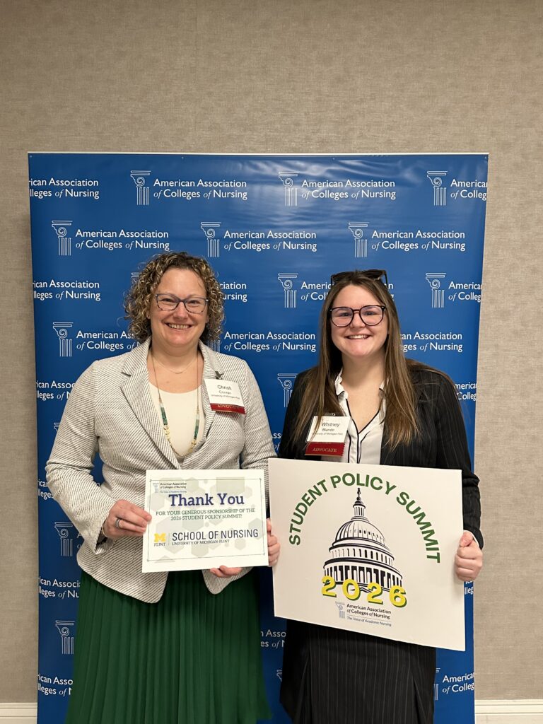 Two women standing in front of an American Association of Colleges of Nursing backdrop holding signs that say "Thank You for your generous sponsorship of the 2026 Student Policy Summit! School of Nursing University of Michigan-Flint" and "Student Policy Summit 2026" with an illustration of the U.S. Capitol building.