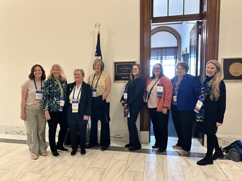 Group of eight women standing in a hallway near a door with a sign that reads "Senator Elissa Slotkin Michigan SR-291" and an American flag.