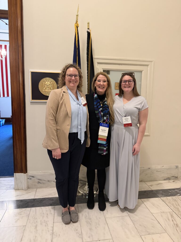 Three women standing together indoors in front of two flags and a framed seal on the wall.