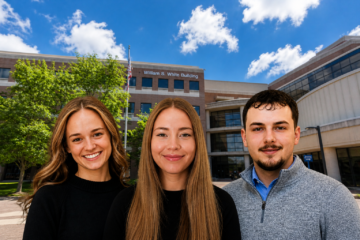 Three people standing outside the William S. White Building on a sunny day with a blue sky and scattered clouds.