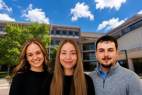 Three people standing outside the William S. White Building on a sunny day with a blue sky and scattered clouds.
