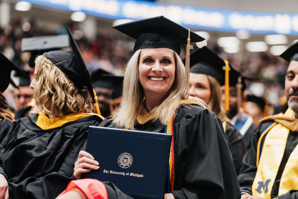A graduate in cap and gown holding a University of Michigan diploma at a commencement ceremony.