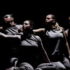 Four dancers in gray sleeveless tops and pants perform a synchronized pose with arms extended against a dark background.
