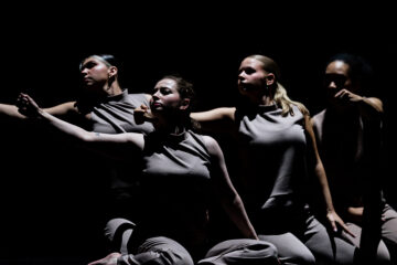 Four dancers in gray sleeveless tops and pants perform a synchronized pose with arms extended against a dark background.