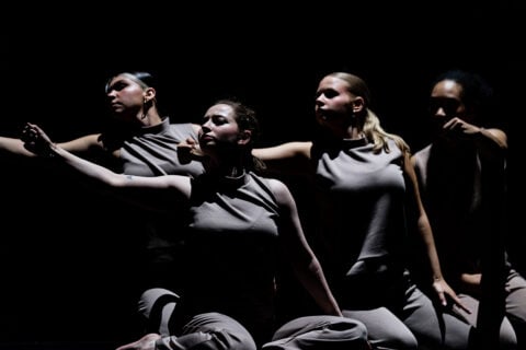 Four dancers in gray sleeveless tops and pants perform a synchronized pose with arms extended against a dark background.