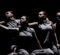 Four dancers in gray sleeveless tops and pants perform a synchronized pose with arms extended against a dark background.