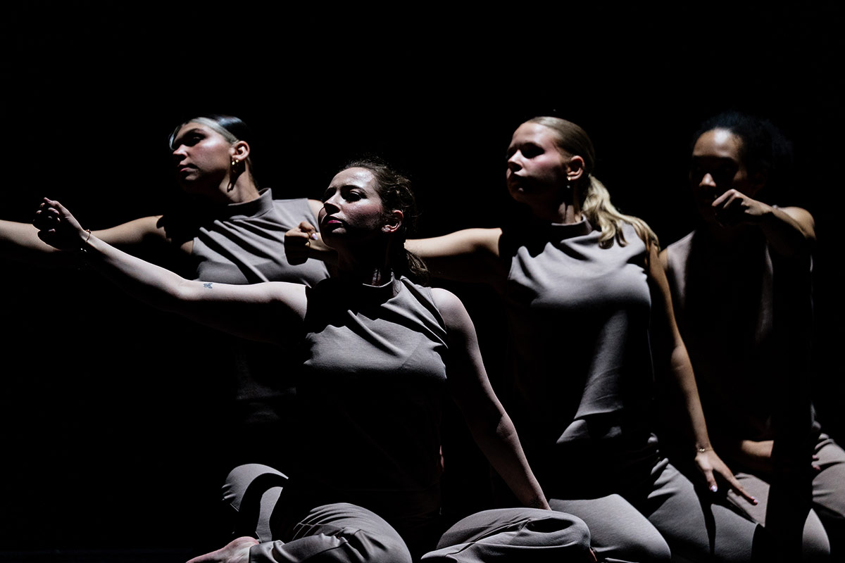 Four dancers in gray sleeveless tops and pants perform a synchronized pose with arms extended against a dark background.