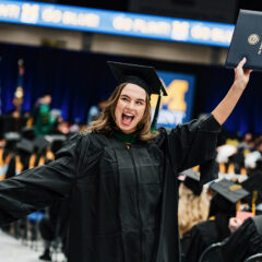 Graduate in black cap and gown holding diploma and a card at a University of Michigan graduation ceremony.