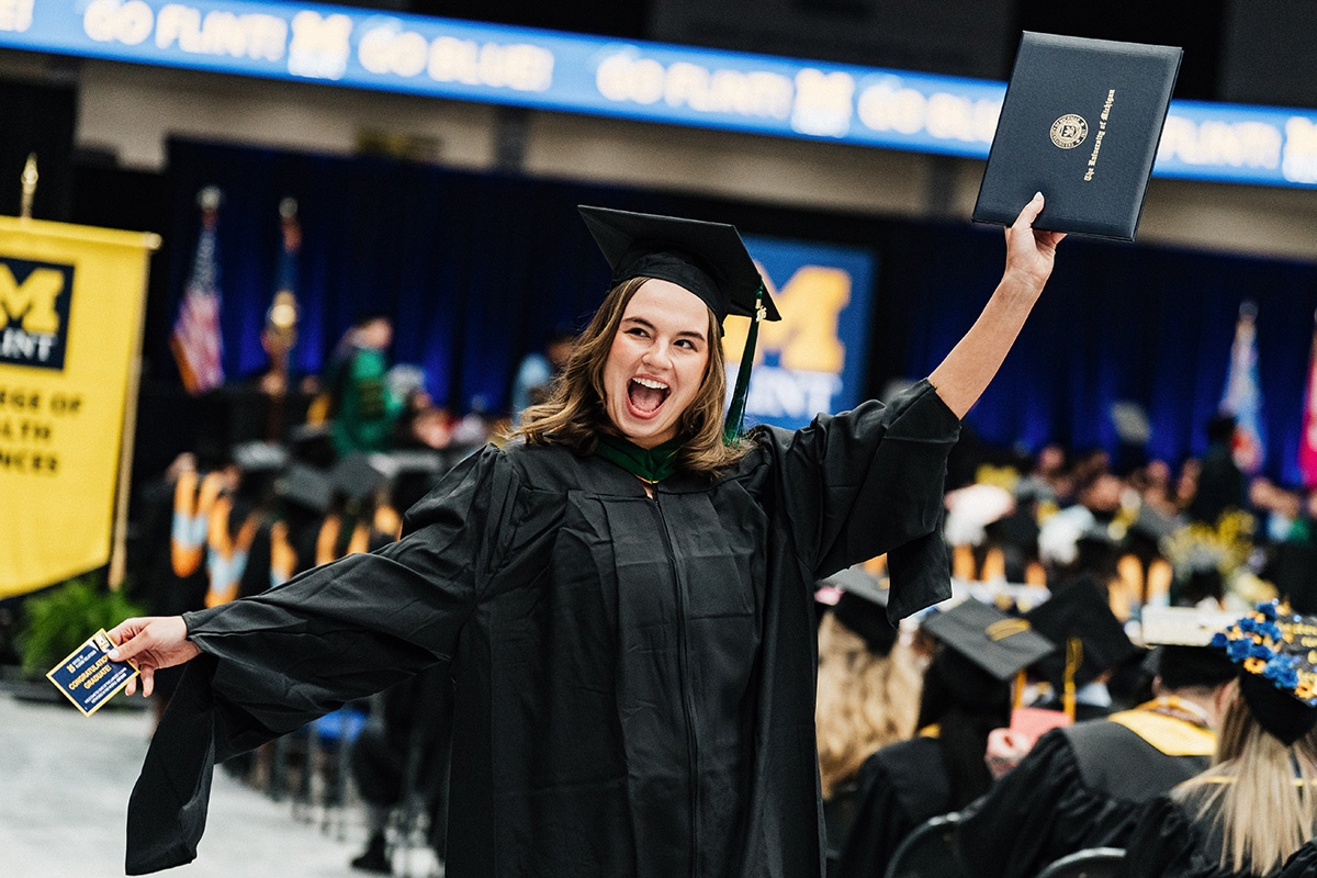 Graduate in black cap and gown holding diploma and a card at a University of Michigan graduation ceremony.
