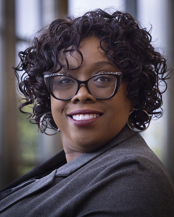 Person with curly dark hair wearing a gray blazer, photographed indoors with blurred background.
