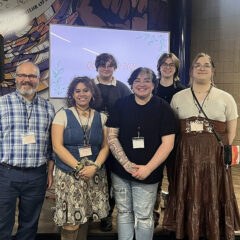 A group of six people standing together in front of a decorative wall and a screen displaying "QUEER 2026."