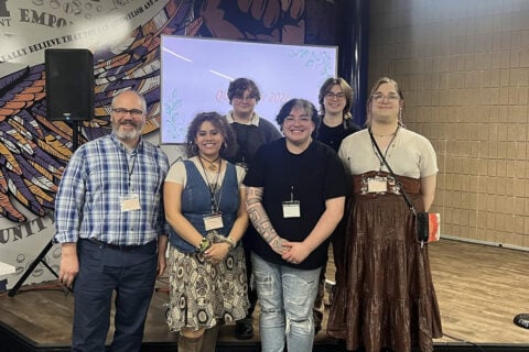 A group of six people standing together in front of a decorative wall and a screen displaying "QUEER 2026."