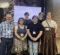 A group of six people standing together in front of a decorative wall and a screen displaying "QUEER 2026."