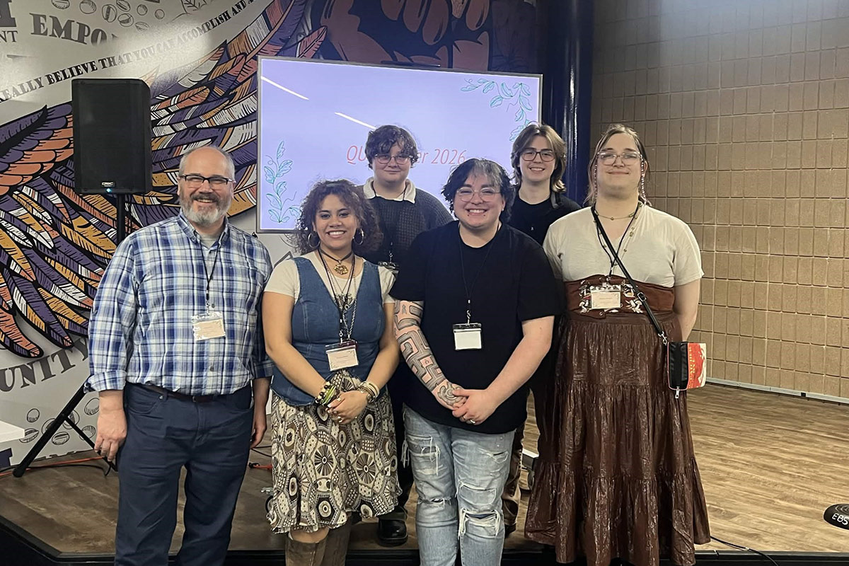 A group of six people standing together in front of a decorative wall and a screen displaying "QUEER 2026."
