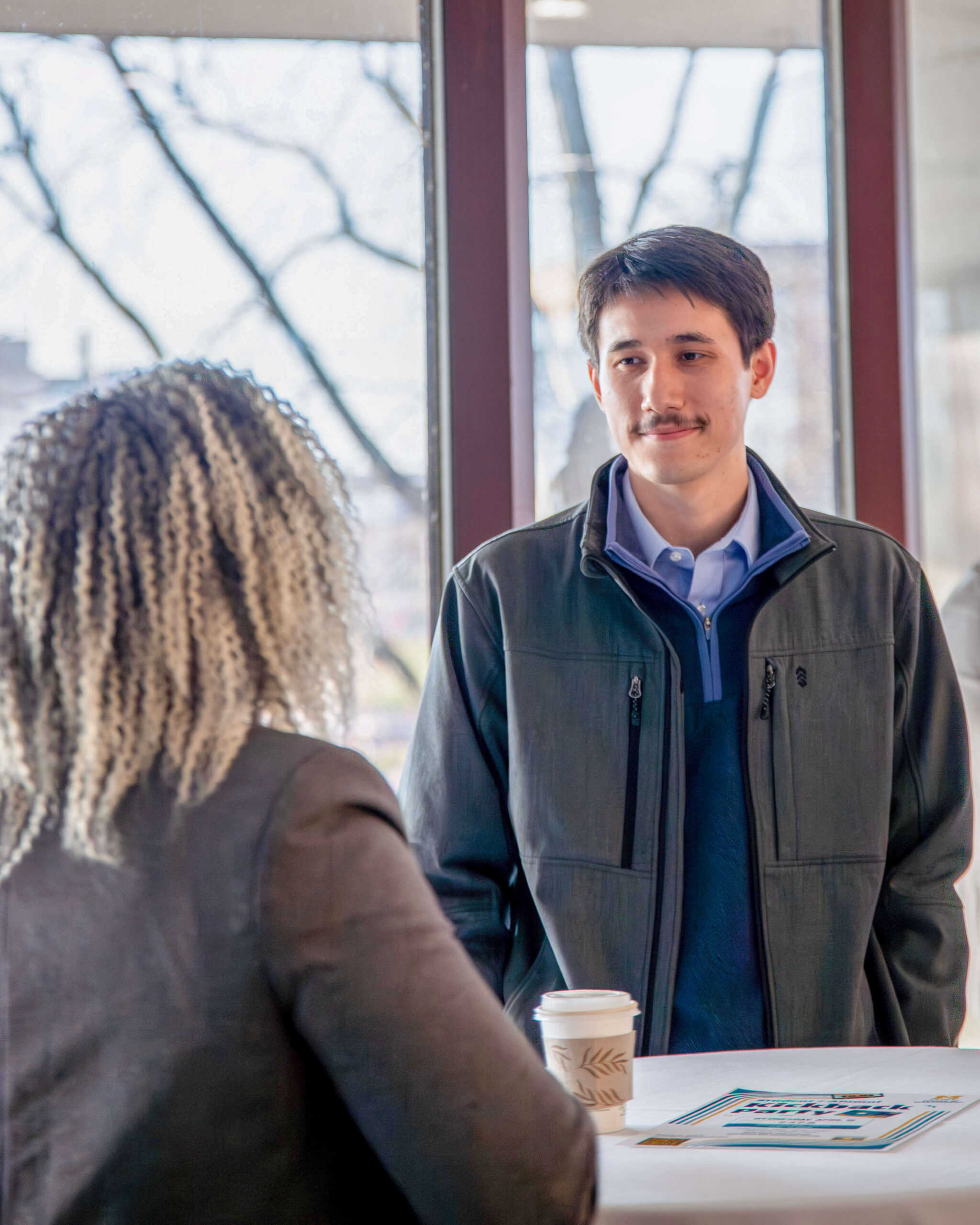 A woman with curly hair is standing at a table facing a man. The man is looking at her and wearing a gray jacket and blue shirt. A coffee cup and a flyer are on the table.