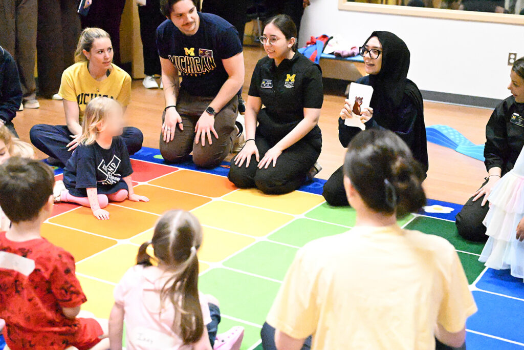 A group of children and adults are gathered on a colorful mat, engaging in an activity. Some adults are kneeling while children sit in front of them, focused on the interaction. One adult holds a card with an image.