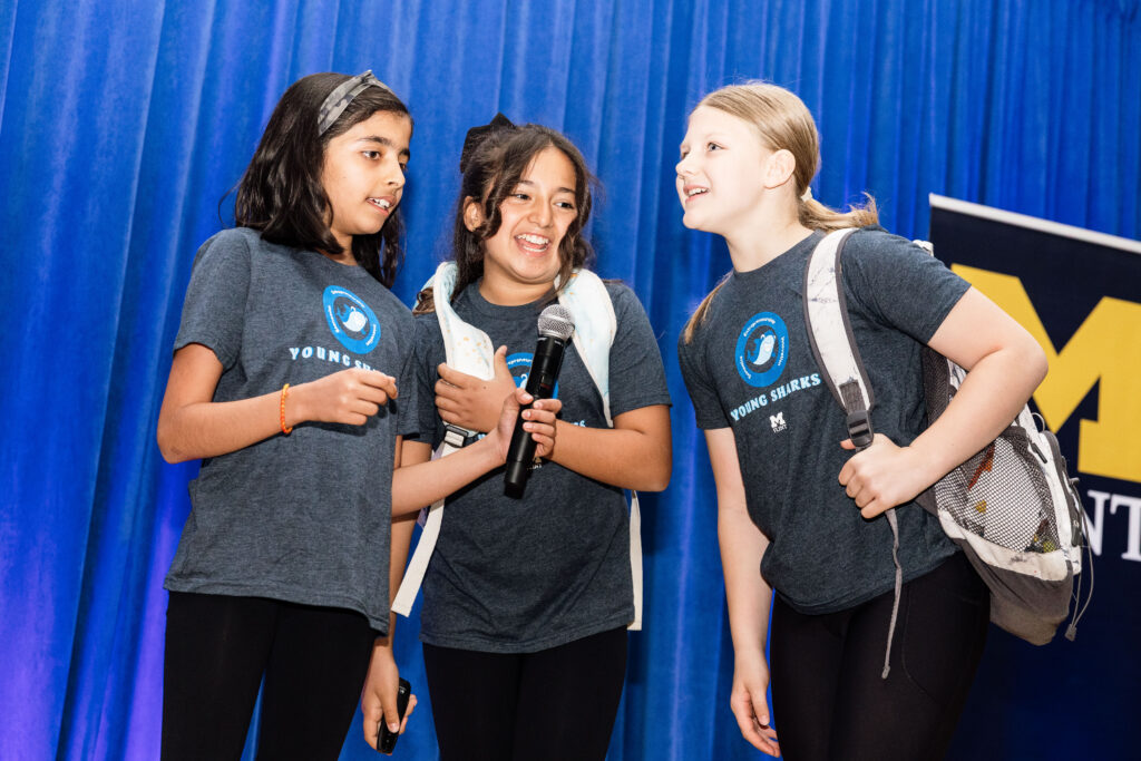 A group of three girls speaking into a microphone on stage wearing backpacks. 