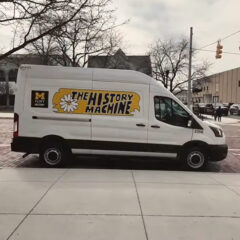 A white van with the text "THE HISTORY MACHINE" and a flower graphic, parked on a brick street near a building.