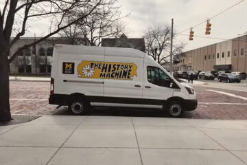 A white van with the text "THE HISTORY MACHINE" and a flower graphic, parked on a brick street near a building.