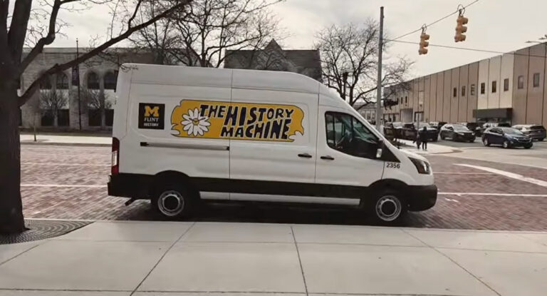 A white van with the text "THE HISTORY MACHINE" and a flower graphic, parked on a brick street near a building.