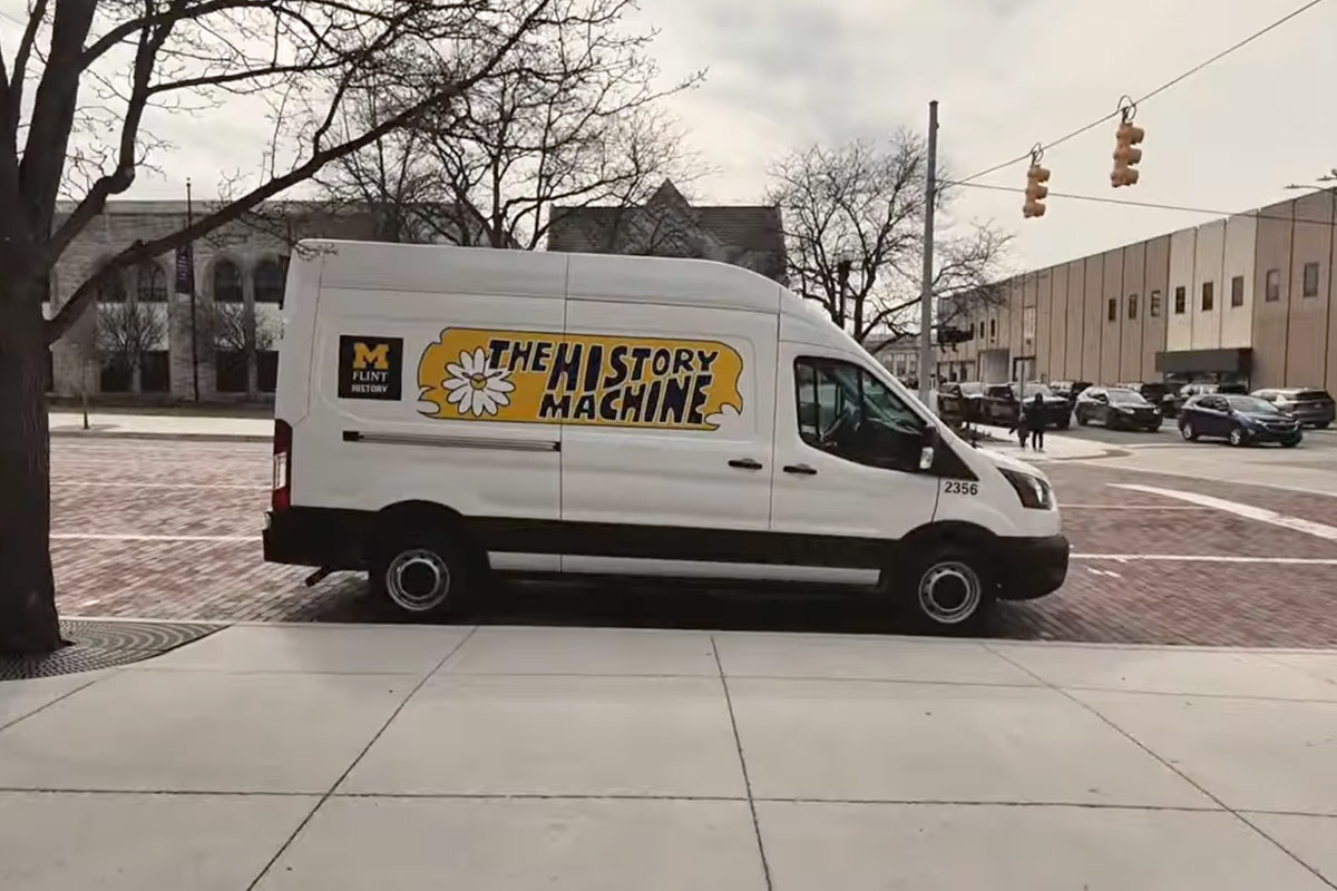 A white van with the text "THE HISTORY MACHINE" and a flower graphic, parked on a brick street near a building.