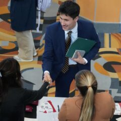 A young man in a suit shakes hands with a woman standing at a table while another woman observes. The setting is a college career fair with patterned carpet that is different shades of tan, orange and blue.