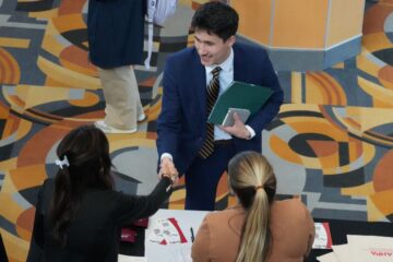 A young man in a suit shakes hands with a woman standing at a table while another woman observes. The setting is a college career fair with patterned carpet that is different shades of tan, orange and blue.