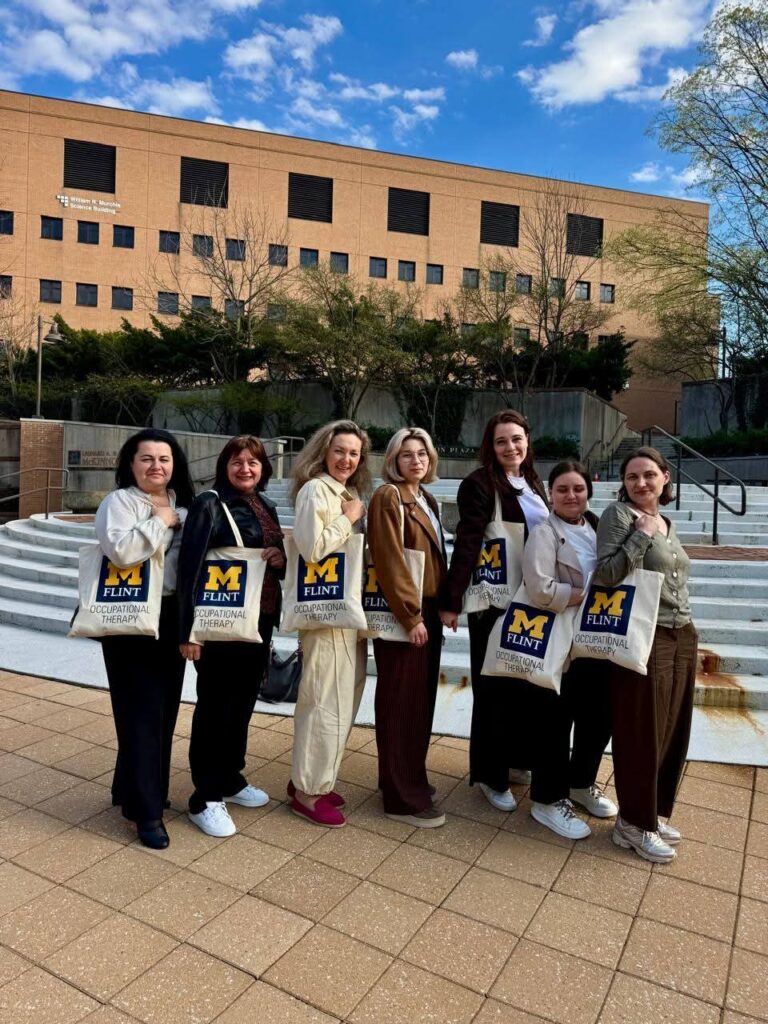 A group of seven individuals stands outdoors, each holding a tote bag with "M FLINT OCCUPATIONAL THERAPY" printed on it, in front of a building.