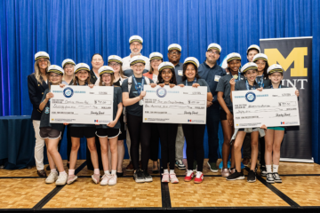 A group of young participants wearing hats stands together, holding oversized checks for various amounts, celebrating their achievements at an event. A banner with the "M" logo is visible in the background.