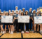 A group of young participants wearing hats stands together, holding oversized checks for various amounts, celebrating their achievements at an event. A banner with the "M" logo is visible in the background.