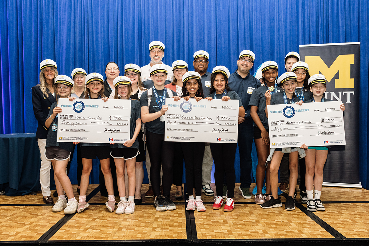 A group of young participants wearing hats stands together, holding oversized checks for various amounts, celebrating their achievements at an event. A banner with the "M" logo is visible in the background.