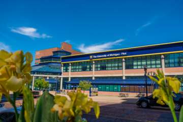The image shows the exterior of the University of Michigan - Flint building, with a clear blue sky and yellow flowers in the foreground.