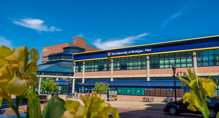The image shows the exterior of the University of Michigan - Flint building, with a clear blue sky and yellow flowers in the foreground.