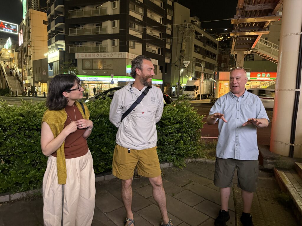 Three people are standing outdoors at night, engaged in conversation. They are surrounded by greenery and urban buildings, with bright signs in the background.