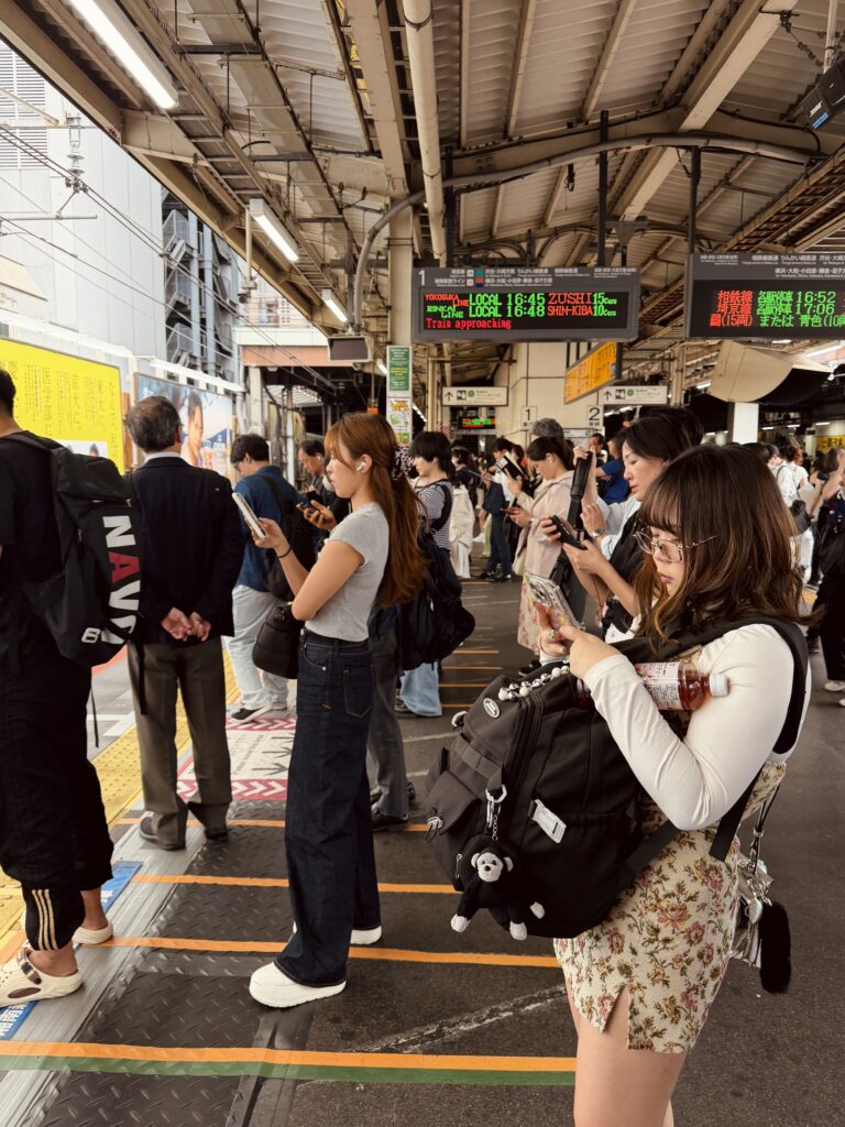 A crowded train station platform with people waiting for a train. A digital display shows train schedules, including "Train approaching."