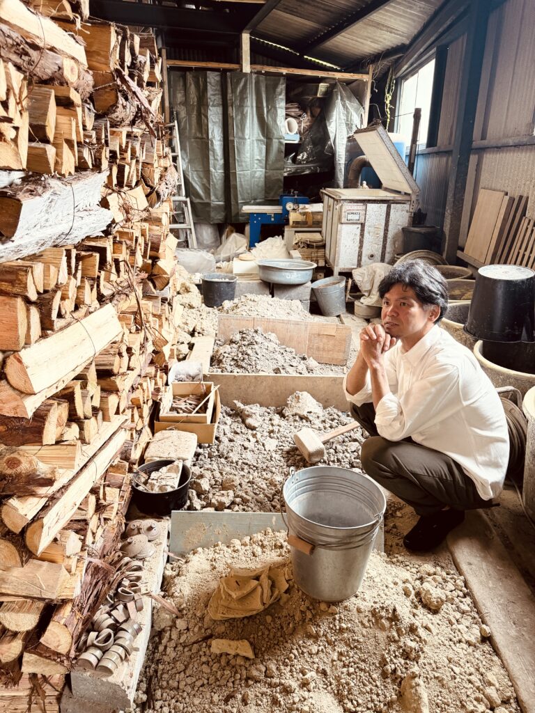 A person in a white shirt sits on the ground in a workshop, surrounded by stacks of firewood, tools, and various materials.