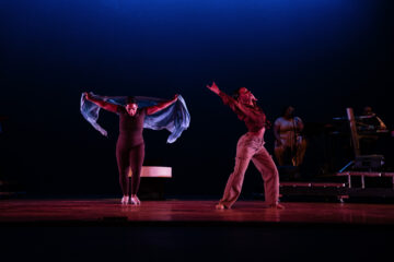 Two dancers perform on stage; one is wearing a black outfit and holding a blue scarf, while the other wears a red shirt and beige pants, striking a dramatic pose. In the background, musicians are visible.