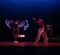Two dancers perform on stage; one is wearing a black outfit and holding a blue scarf, while the other wears a red shirt and beige pants, striking a dramatic pose. In the background, musicians are visible.