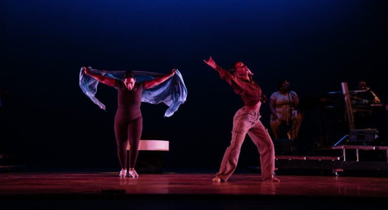 Two dancers perform on stage; one is wearing a black outfit and holding a blue scarf, while the other wears a red shirt and beige pants, striking a dramatic pose. In the background, musicians are visible.