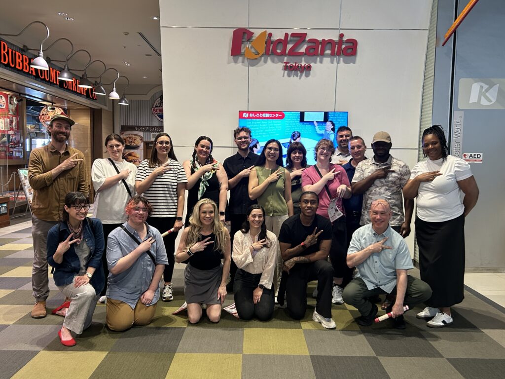 A diverse group of 18 people poses together in front of a "KidZania Tokyo" sign, making hand gestures.