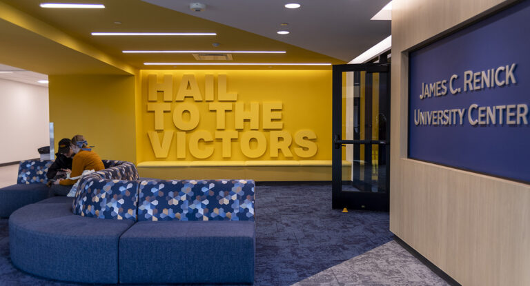 Interior of the James C. Renick University Center featuring a large yellow wall with the text "HAIL TO THE VICTORS" and a blue seating area with patterned cushions.