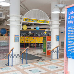 Entrance to the University Pavilion featuring a sign that reads "UNIVERSITY PAVILION," colorful hanging lanterns, and informational displays about UM-Flint and the city of Flint.