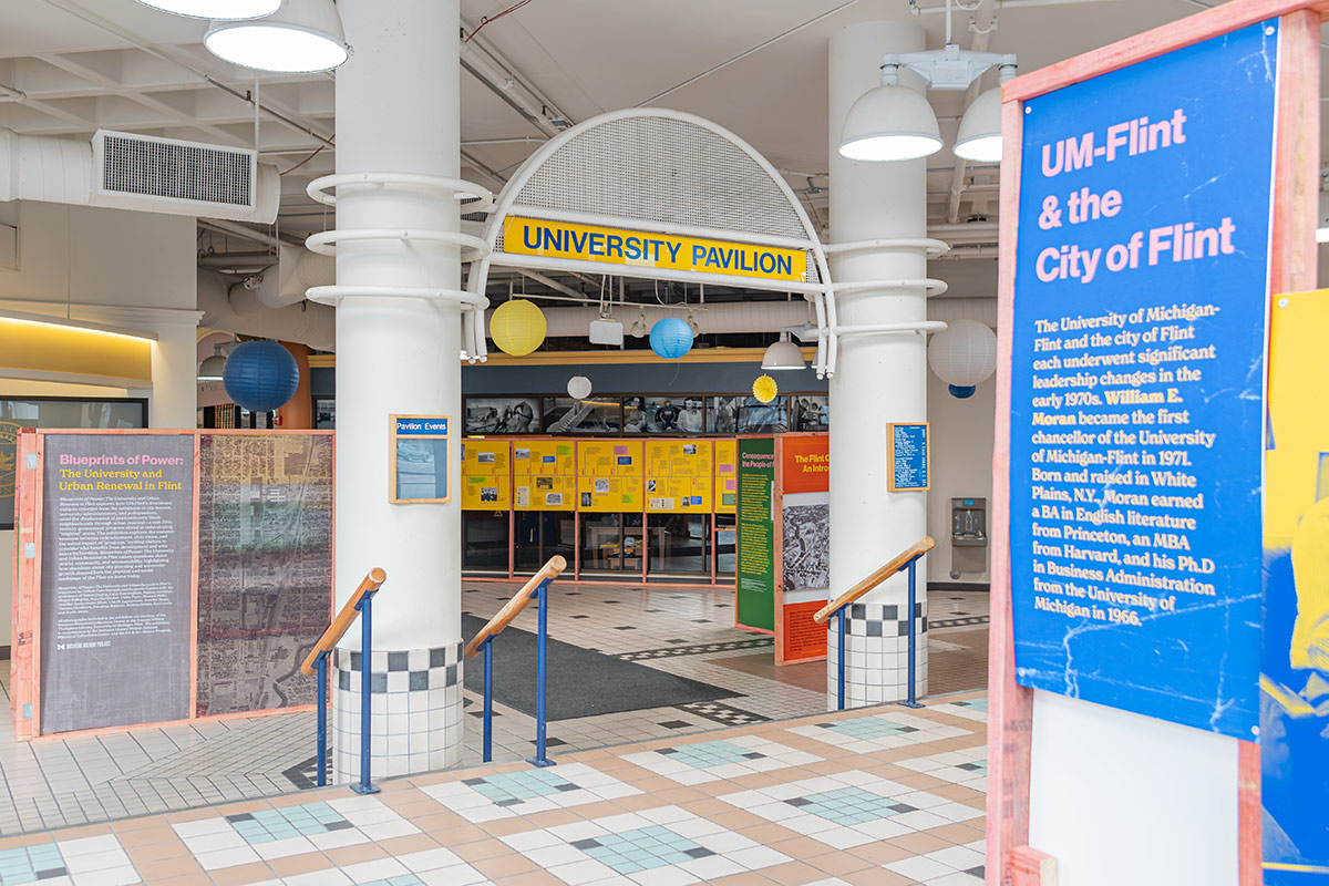 Entrance to the University Pavilion featuring a sign that reads "UNIVERSITY PAVILION," colorful hanging lanterns, and informational displays about UM-Flint and the city of Flint.
