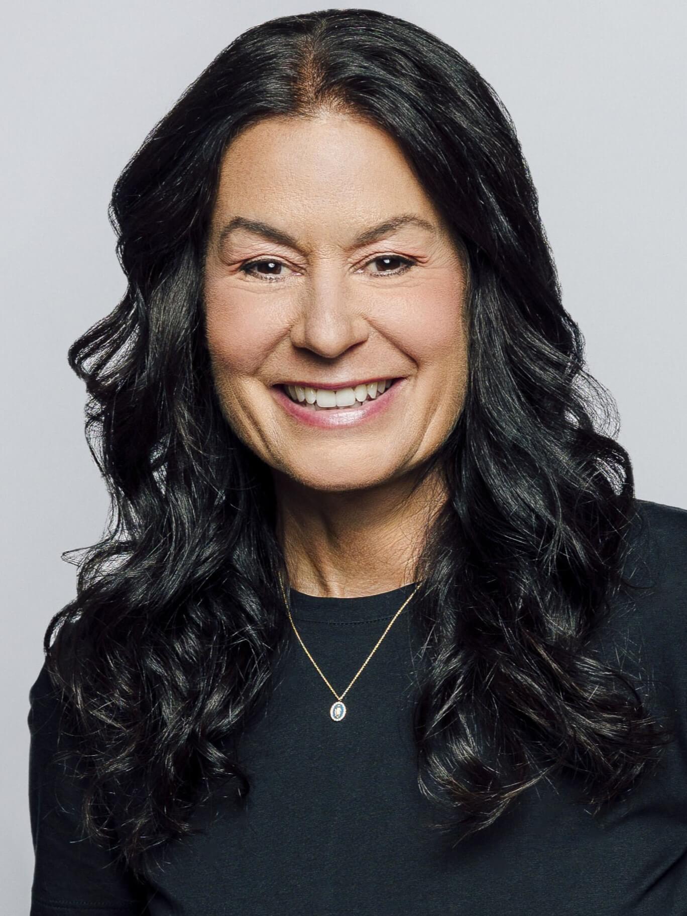 A professional portrait of a woman smiling warmly. She has long, wavy black hair and is wearing a black t-shirt with a delicate gold necklace and small pendant. The background is a neutral light gray.
