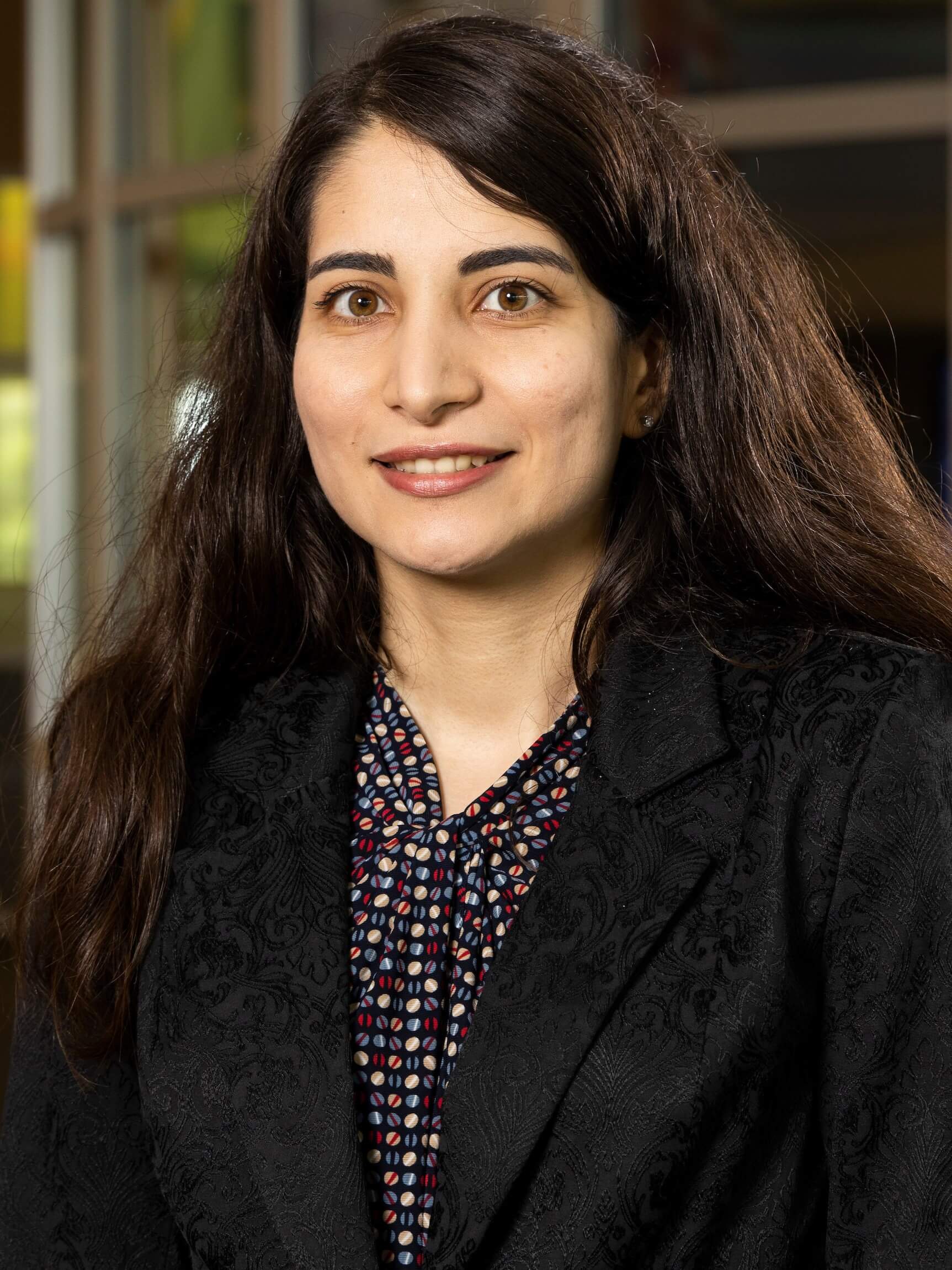 A woman with long, dark hair is smiling. She is wearing a black blazer over a patterned blouse while standing in an indoor setting with soft lighting.