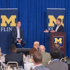 A master of ceremonies gestures while addressing a guest lecturer seated on stage to the left. Two banners displaying the University of Michigan-Flint logo are in the background.